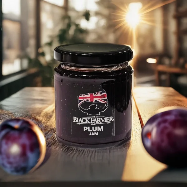 A jar of The Black Farmer Plum Jam on a rustic wooden table, surrounded by fresh plums, with warm sunlight streaming through the background.