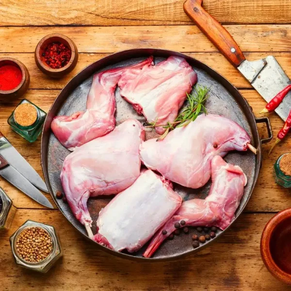 Raw jointed rabbit pieces arranged in a pan, surrounded by spices, herbs, and cooking utensils on a rustic wooden surface, ready for preparation.