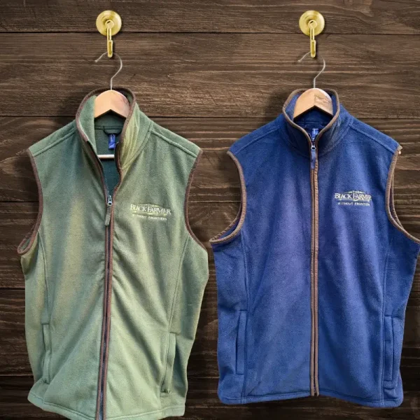 The Black Farmer gilet available in moss green and navy blue, featuring a sleeveless design with an embroidered logo, displayed on wooden hangers.